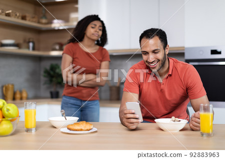 Incredulous sad millennial african american wife looks at smartphone of laughing husband in kitchen interior Incredulous sad millennial african american wife looks at smartphone of laughing husband in kitchen interior 92883963