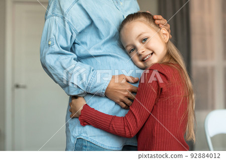 Pretty little girl hugging her pregnant mother big belly and smiling at camera, kid bonding with expecting mom 92884012