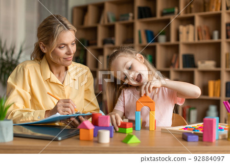 Child psychology exam. Little girl playing with colorful wooden blocks, professional kids psychologist writing notes 92884097