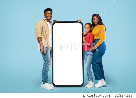 Young black family posing near huge smartphone with blank screen, advertising mobile app or website, mockup Young black family posing near huge smartphone with blank screen, advertising mobile app or website, mockup 92884158