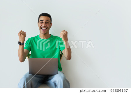 Overjoyed arab guy making YES gesture, sitting on chair with laptop, celebrating success or achievement over white wall 92884167