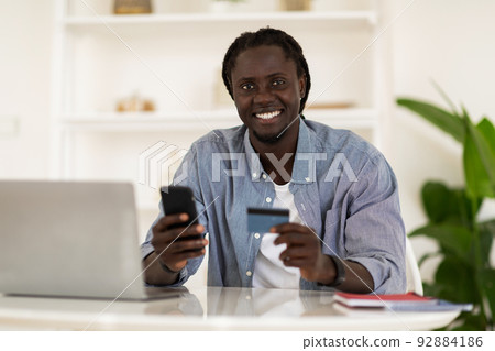 Online Payments. Smiling Black Male Freelancer Using Smartphone And Credit Card 92884186