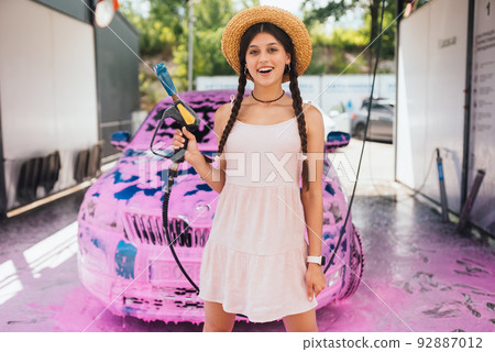 Young woman hose stands in front of a car covered in pink foam Young woman hose stands in front of a car covered in pink foam 92887012