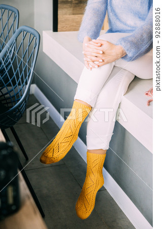 Comforting cozy woman in knitted woolen winter yellow socks, white trousers and blue warm sweater resting sitting on a bench at cafe in winter time. Cozy time Comforting cozy woman in knitted woolen winter yellow socks, white trousers and blue warm sweater resting sitting on a bench at cafe in winter time. Cozy time 92888016