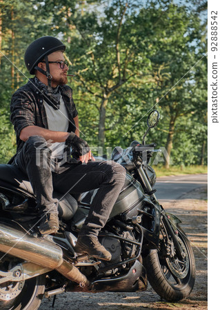 Young male biker in helmet travels on a motorcycle alone, stopped and smoking on the side of a forest road 92888542