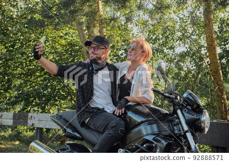 Middle age couple riding a motorcycle having fun and taking a selfie on a mobile phone camera Middle age couple riding a motorcycle having fun and taking a selfie on a mobile phone camera 92888571