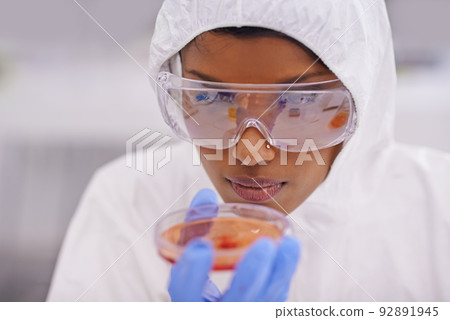 Careful observation. A young scientist in protective clothing examining a petri dish in her lab. 92891945