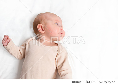 Newborn baby in beige bodysuit looks aside lying on bed Newborn baby in beige bodysuit looks aside lying on bed 92892072