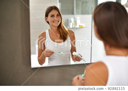 Step one to a brilliant smile. Over the shoulder portrait of an attractive young woman brushing her teeth in a bathroom mirror. 92892115