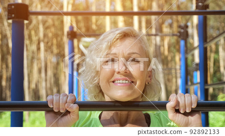 Mature woman does pull-ups smiling on sports ground closeup Mature woman does pull-ups smiling on sports ground closeup 92892133