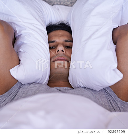 No-one can sleep with all this noise. A young man covering his ears with his pillow. 92892209