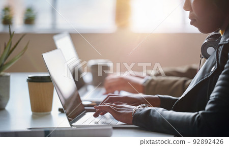 A focused young businesswoman typing on her laptop and wearing headphones. A young african american businesswoman working on her laptop next to a colleague. A businesswoman typing emails on her laptop 92892426