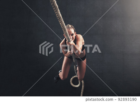 Hold on tight through your challenges. Shot of a sporty young woman climbing a rope in a gym. 92893190
