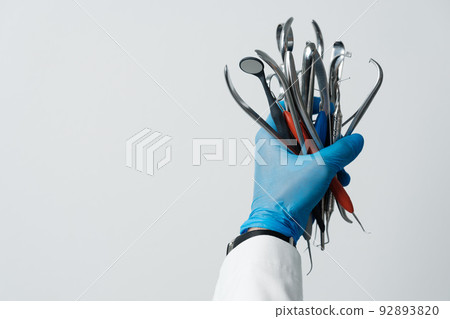 The dentist's hand in a blue glove holds bunch of dental instruments, dental mirror, tweezers, probe hooks. The dentist's hand in a blue glove holds bunch of dental instruments, dental mirror, tweezers, probe hooks. 92893820