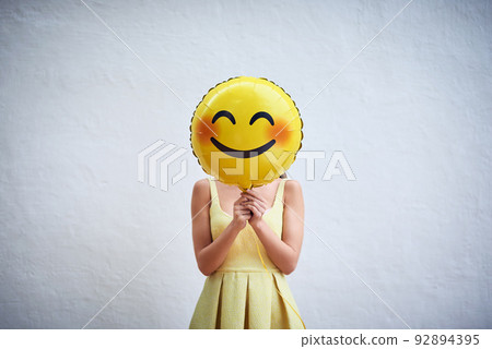 Hold on to your smile and spread the happiness. Studio shot of a young woman holding a balloon with a smiley in front of her face. 92894395