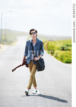 All I need is my guitar and the open road. Trendy young male wearing sunglasses standing in the middle of the road holding a guitar. 92894722