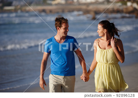 Hand in hand and face to face. Shot of an affectionate couple walking on the beach. 92896128