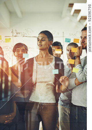 Group of marketing professionals or designers brainstorming ideas, writing on transparent board with sticky notes during meeting. Diverse team of colleagues discussing work project schedule plan. 92896798