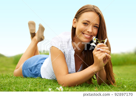 Cute and fresh-faced beauty. Cute young woman smiling and holding a flower while lying on the grass. 92897170