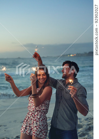 happy couple holding sparklers celebrating new years eve on beach at sunset with sparkle firework 92902027