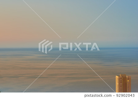 Calm waters during sunset with a hotel building overlooking the ocean. Aerial view of the sea during dusk. Copy space of small waves on calm water in the sea, bright afterglow in a clear summer sky Calm waters during sunset with a hotel building overlooking the ocean. Aerial view of the sea during dusk. Copy space of small waves on calm water in the sea, bright afterglow in a clear summer sky 92902043