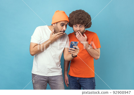 Portrait of two attractive shocked young adult hipster men standing, using smartphone, reading breaking news in social networks, covering open mouth. Indoor studio shot isolated on blue background. 92908947