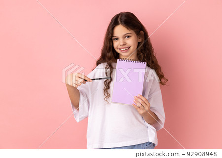Portrait of beautiful little girl wearing white T-shirt pointing at paper notebook, looking smiling at camera, has positive expression. Indoor studio shot isolated on pink background. Portrait of beautiful little girl wearing white T-shirt pointing at paper notebook, looking smiling at camera, has positive expression. Indoor studio shot isolated on pink background. 92908948