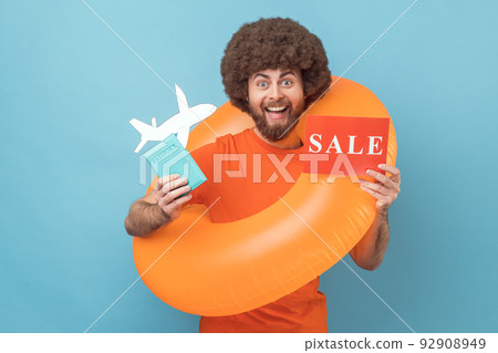 Smiling man with Afro hairstyle wearing orange T-shirt standing with orange rubber ring, holding passport document and sale card, tours for low cost. Indoor studio shot isolated on blue background Smiling man with Afro hairstyle wearing orange T-shirt standing with orange rubber ring, holding passport document and sale card, tours for low cost. Indoor studio shot isolated on blue background 92908949