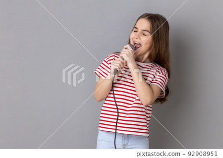 Portrait of positive little girl wearing striped T-shirt singing songs, holding microphone, singer making performance, keeps eyes closed. Indoor studio shot isolated on gray background. Portrait of positive little girl wearing striped T-shirt singing songs, holding microphone, singer making performance, keeps eyes closed. Indoor studio shot isolated on gray background. 92908951