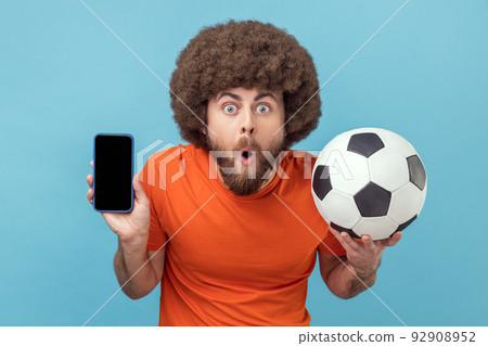 Portrait of shocked man with Afro hairstyle wearing T-shirt standing with soccer ball and showing cell phone with empty display for advertisement. Indoor studio shot isolated on blue background. 92908952