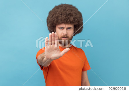 Portrait of man with Afro hairstyle in orange T-shirt making stop gesture showing palm of hand, conflict prohibition warning about danger, stop bullying. Indoor studio shot isolated on blue background 92909016