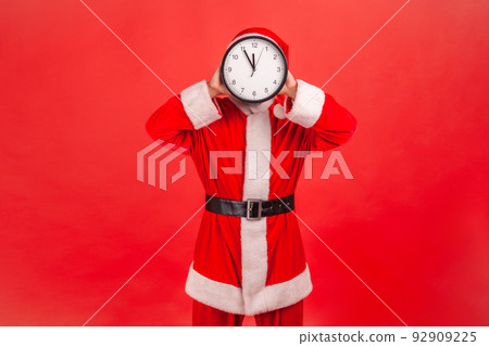 Portrait of unknown man in santa claus costume standing covering face with wall clock, wasting his time, procrastination, organization of working time. Indoor studio shot isolated on red background. Portrait of unknown man in santa claus costume standing covering face with wall clock, wasting his time, procrastination, organization of working time. Indoor studio shot isolated on red background. 92909225
