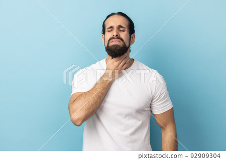 Portrait of unhealthy man with beard wearing white T-shirt suffering from sore throat, keeping hand on her neck, frowning face and closed eyes. Indoor studio shot isolated on blue background. Portrait of unhealthy man with beard wearing white T-shirt suffering from sore throat, keeping hand on her neck, frowning face and closed eyes. Indoor studio shot isolated on blue background. 92909304