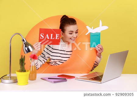 Optimistic woman with rubber ring, holding card with sale inscription, passport and paper plane, sales for tours, video call. Indoor studio studio shot isolated on yellow background. Optimistic woman with rubber ring, holding card with sale inscription, passport and paper plane, sales for tours, video call. Indoor studio studio shot isolated on yellow background. 92909305