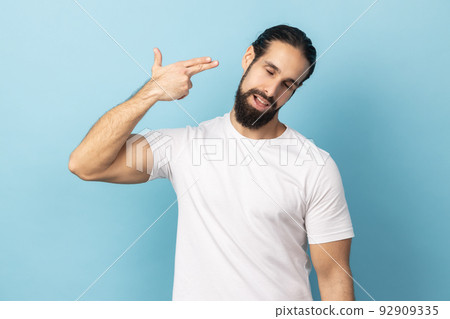 Headshot, kill me please. Depressed man with beard wearing white T-shirt committing suicide with finger gun gesture, desperate face expression. Indoor studio shot isolated on blue background. Headshot, kill me please. Depressed man with beard wearing white T-shirt committing suicide with finger gun gesture, desperate face expression. Indoor studio shot isolated on blue background. 92909335