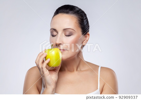Closeup portrait of beautiful charming middle aged woman holding smelling green apple, vitamins for beauty and skin care, keeps eyes closed. Indoor studio shot isolated on gray background. Closeup portrait of beautiful charming middle aged woman holding smelling green apple, vitamins for beauty and skin care, keeps eyes closed. Indoor studio shot isolated on gray background. 92909337