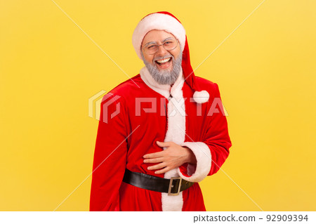 Portrait of elderly man with gray beard wearing santa claus costume holding his belly and laughing out loud, hearing funny story or joke. Indoor studio shot isolated on yellow background. 92909394