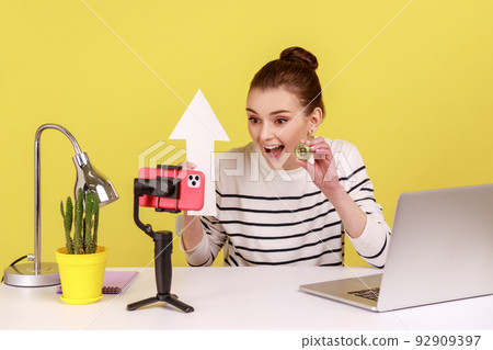 Woman blogger holding golden bitcoin and white arrow pointing up, posing at smartphone camera, talking about way to make money, e-commerce. Indoor studio studio shot isolated on yellow background. Woman blogger holding golden bitcoin and white arrow pointing up, posing at smartphone camera, talking about way to make money, e-commerce. Indoor studio studio shot isolated on yellow background. 92909397