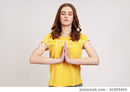 Young teenager girl practicing yoga, holding hands in namaste and keeping her eyes closed, meditating, praying for peace and love, looks calm. Indoor studio shot isolated on gray background. 92909404