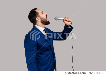Side view of bearded man singing songs, holding microphone, singer making performance, keeps eyes closed, wearing official style suit. Indoor studio shot isolated on gray background. 92909416
