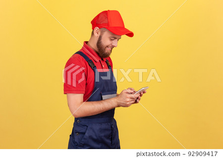 Side view of smiling worker man typing on mobile phone, using cellphone messenger to accept online order as delivery, repair and maintenance services. Indoor studio shot isolated on yellow background. 92909417