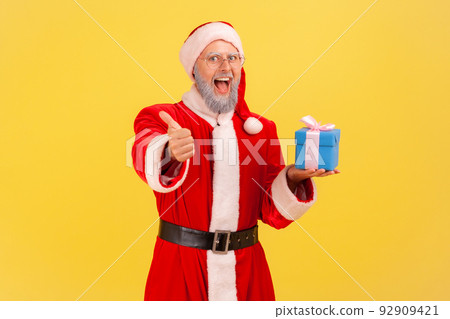 Excited positive elderly man with gray beard wearing santa claus costume with blue wrapped present box. showing thumb up to camera with toothy smile. Indoor studio shot isolated on yellow background. 92909421