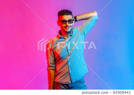 Portrait of man in shirt and glasses standing with raised hand and looking at camera with positive smile and optimistic expression. Indoor studio shot isolated on colorful neon light background. 92909441