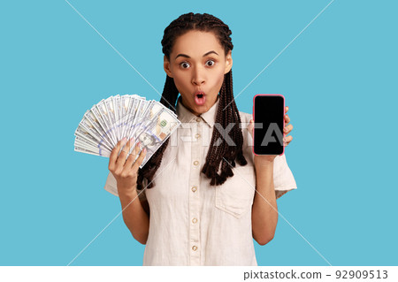 Attractive shocked woman with black dreadlocks holding fan of dollar banknotes and smart phone with empty screen, making money online. Indoor studio shot isolated on blue background. Attractive shocked woman with black dreadlocks holding fan of dollar banknotes and smart phone with empty screen, making money online. Indoor studio shot isolated on blue background. 92909513