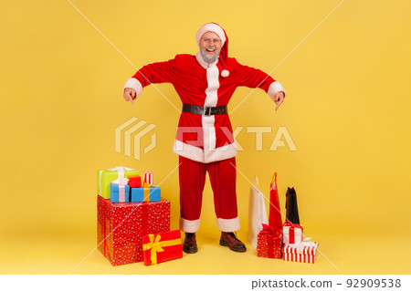Full length portrait of smiling elderly man with gray beard wearing santa claus costume pointing at many present boxes for Christmas holidays. Indoor studio shot isolated on yellow background. 92909538