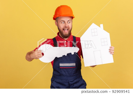 Portrait of excited worker man holding big paper key and house in hands, yelling something with excitement, wearing overalls and red cap. Indoor studio shot isolated on yellow background. Portrait of excited worker man holding big paper key and house in hands, yelling something with excitement, wearing overalls and red cap. Indoor studio shot isolated on yellow background. 92909541