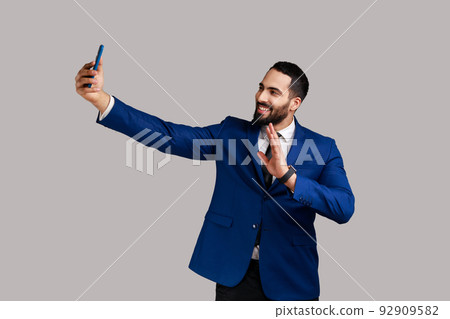 Man making selfie on smartphone camera, blogger communicating, recording video for followers, waving hand, saying hello, wearing official style suit. Indoor studio shot isolated on gray background. 92909582
