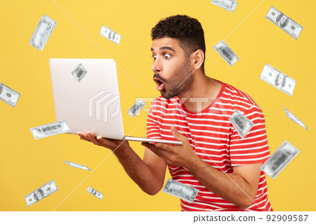 Money falling and shocked rich man. Money rain and portrait of shocked man holding laptop and looking at laptop display with big shocked unbelievable eyes. Indoor isolated on yellow background Money falling and shocked rich man. Money rain and portrait of shocked man holding laptop and looking at laptop display with big shocked unbelievable eyes. Indoor isolated on yellow background 92909587