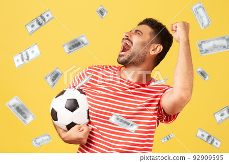 Money rain and rich lucky man extremely happy, holding soccer ball, celebrating victory of favourite football soccer team and his betting. Indoor studio shot isolated on yellow background 92909595