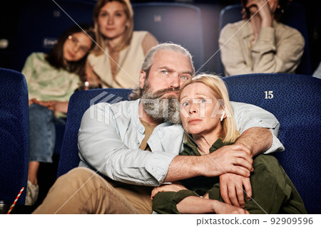 Mature adult Caucasian man with beard on face hugging his wife while watching sad drama film at cinema 92909596